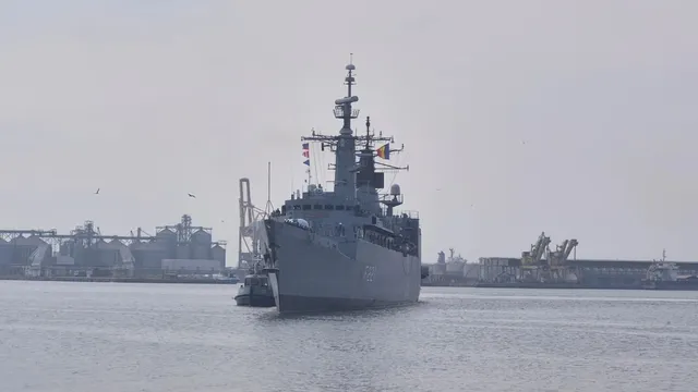 O navă militară gri navighează pe apă, având structuri portuare și industriale în fundal sub un cer înnorat.