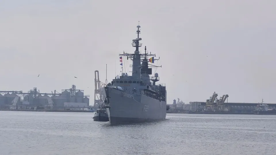 O navă militară gri navighează pe apă, având structuri portuare și industriale în fundal sub un cer înnorat.