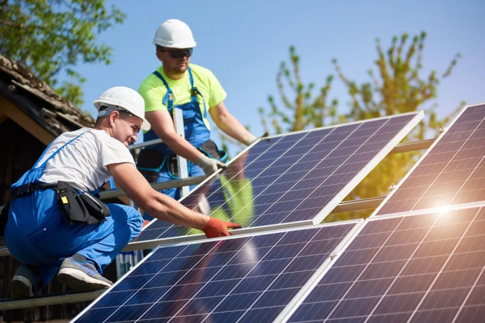 Doi bărbați cu căști albe și haine de lucru instalează panouri solare pe o structură metalică sub cerul senin.