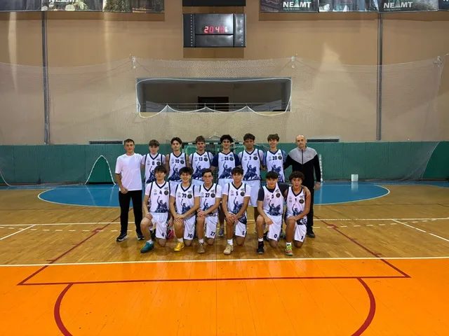 O echipă de baschet formată din unsprezece jucători și un antrenor pozează pe un teren de baschet în interiorul unei săli de sport.