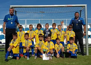 Un grup de copii în echipamente galben și albastru stă așezat pe iarbă în fața unei porți de fotbal, alături de doi adulți.