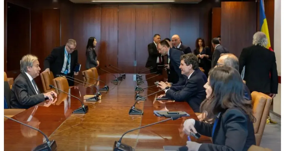 Un grup de persoane stă la o masă lungă de conferințe din lemn într-o încăpere cu pereți din panouri maro.