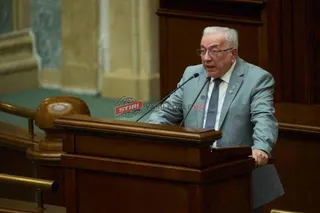 Un bărbat în vârstă, cu ochelari și costum, vorbește la un pupitru cu microfoane, într-o sală de ședințe.