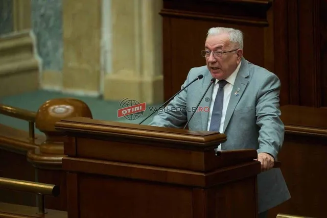 Un bărbat în vârstă, cu ochelari și costum, vorbește la un pupitru cu microfoane, într-o sală de ședințe.