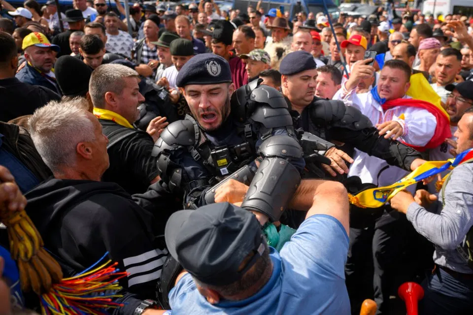 Unii polițiști în echipament de protecție negru intervin într-o mulțime de oameni care poartă haine colorate și steaguri.