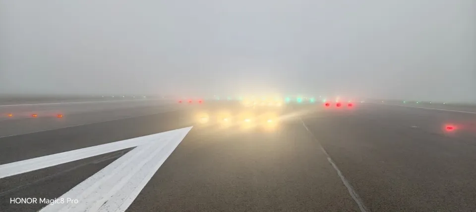 O pistă de aterizare este iluminată de lumini colorate, roșii, verzi și galbene, sub un cer dens și ceață.