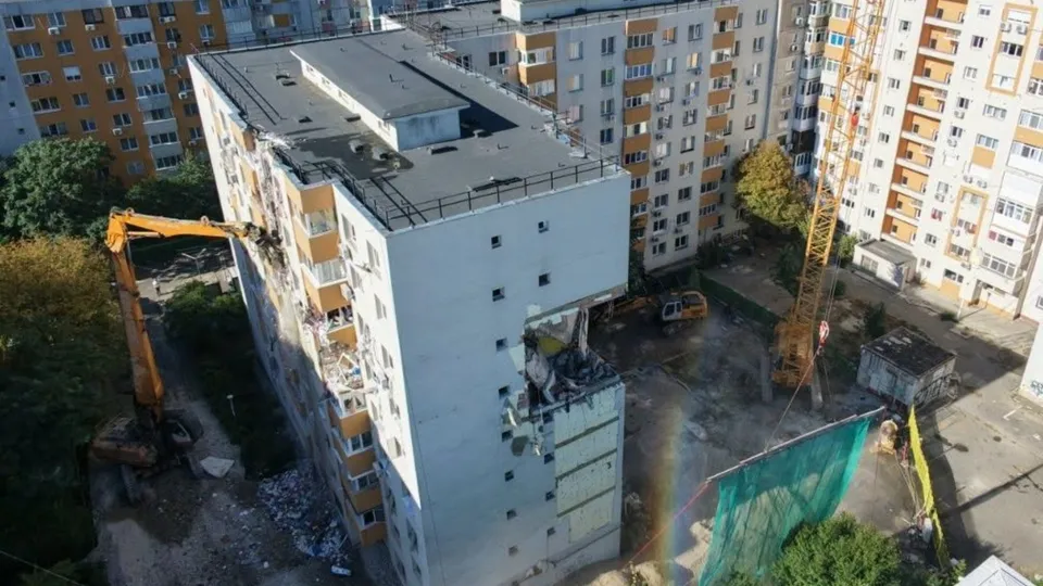 O excavatoră portocalie lucrează lângă un bloc de apartamente cu etajele superioare lipsă și resturi de beton pe sol.