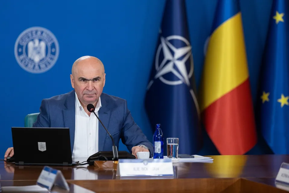 Un bărbat cu sacou albastru și cămașă albă stă la o masă de conferință lângă un laptop, un microfon și un pahar de apă.