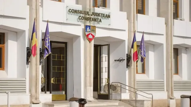 O clădire albă cu ferestre maronii prezintă două steaguri românești și două steaguri albastre afișate pe fațadă lângă intrări.