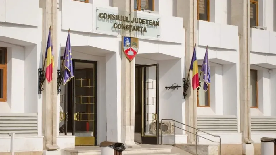 O clădire albă cu ferestre maronii prezintă două steaguri românești și două steaguri albastre afișate pe fațadă lângă intrări.