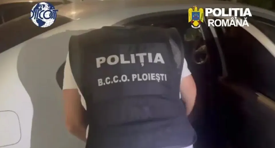 Un polițist din spate purtând o vestă cu inscripția 'POLIȚIA B.C.C.O. PLOIEȘTI' se află lângă o mașină albă de poliție.
