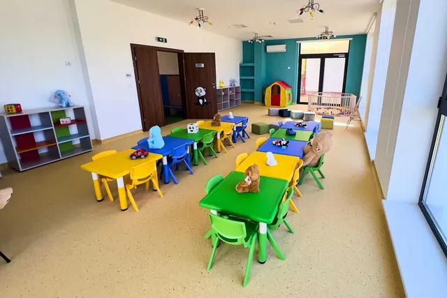 O cameră de joacă cu mese mici colorate, scaune de plastic și jucării împrăștiate pe un podea de culoare deschisă.