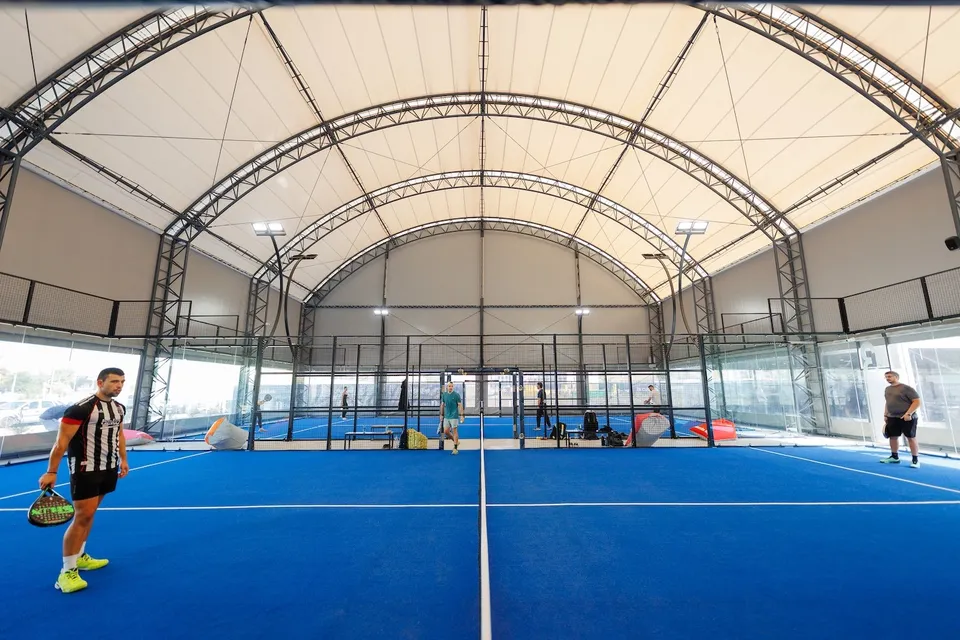 Câțiva bărbați sunt pe terenuri de padel cu suprafață albastră, sub o structură metalică acoperită cu material textil alb.