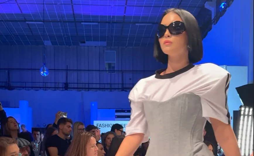 O femeie cu ochelari de soare negri poartă un tricou alb cu detalii negre și o fustă gri, stând în fața unui public.