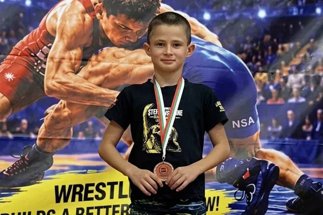 Un băiat cu un medalie la gât stă în fața unei imagini mari cu doi sportivi care practică wrestling.