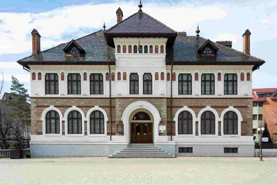 O clădire cu acoperiș înclinat, ferestre arcuate și fațadă albă și maro, situată pe o suprafață pavată deschisă sub un cer înnorat.
