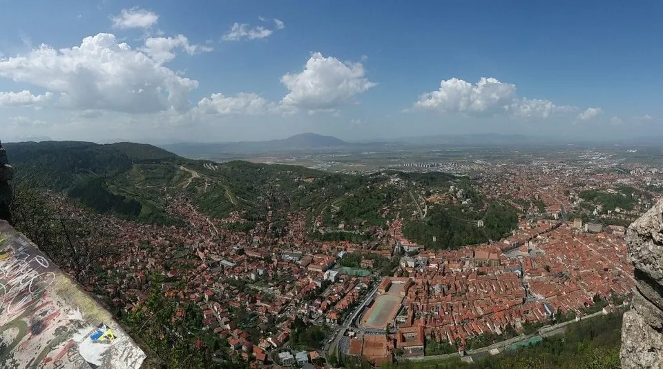 O vedere panoramică de la înălțime asupra unui oraș cu numeroase case cu acoperișuri roșii, situat la poalele unor dealuri verzi sub un cer înnorat.