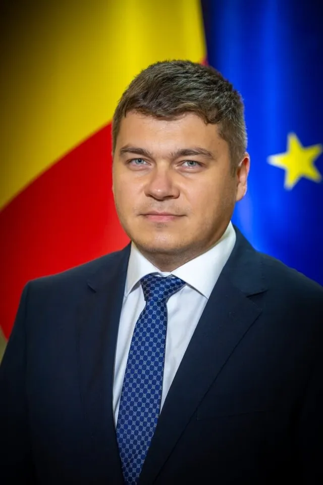 Un bărbat cu părul scurt, purtând sacou închis la culoare, cămașă albă și cravată albastră cu model, stă în fața unor steaguri colorate.