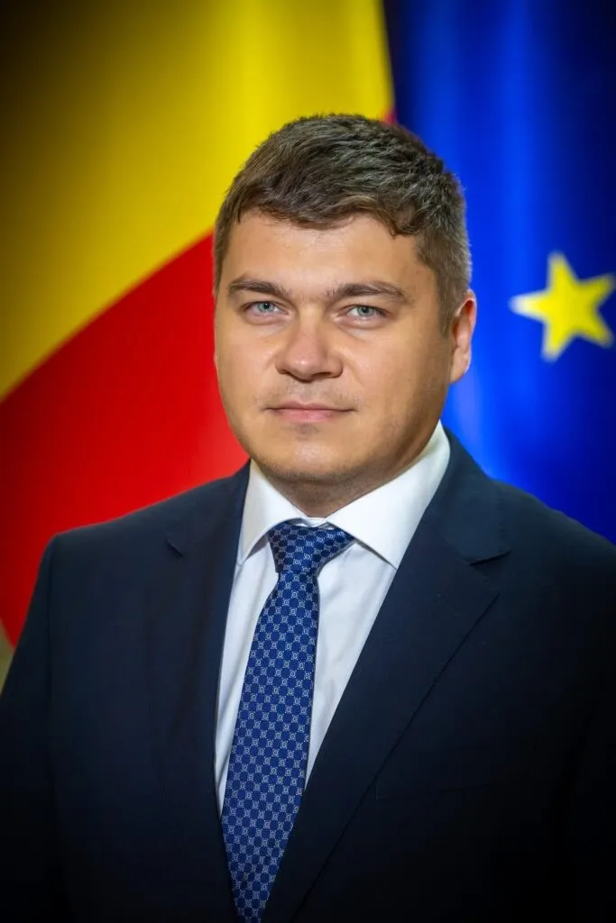 Un bărbat cu părul scurt, purtând sacou închis la culoare, cămașă albă și cravată albastră cu model, stă în fața unor steaguri colorate.