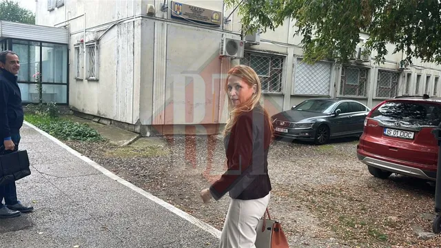 O femeie cu părul blond și o geantă de mână merge pe un drum de pietriș lângă o clădire și câteva mașini parcate.