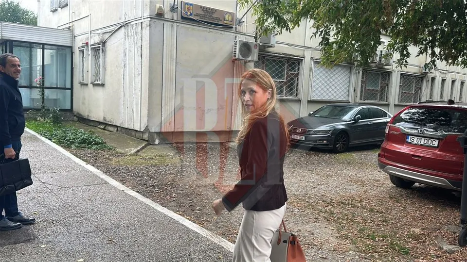 O femeie cu părul blond și o geantă de mână merge pe un drum de pietriș lângă o clădire și câteva mașini parcate.
