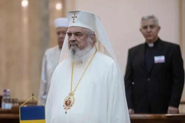 Un bărbat cu barbă albă poartă o vestimentație albă și o pălărie albă cu o cruce, având un medalion suspendat la gât.