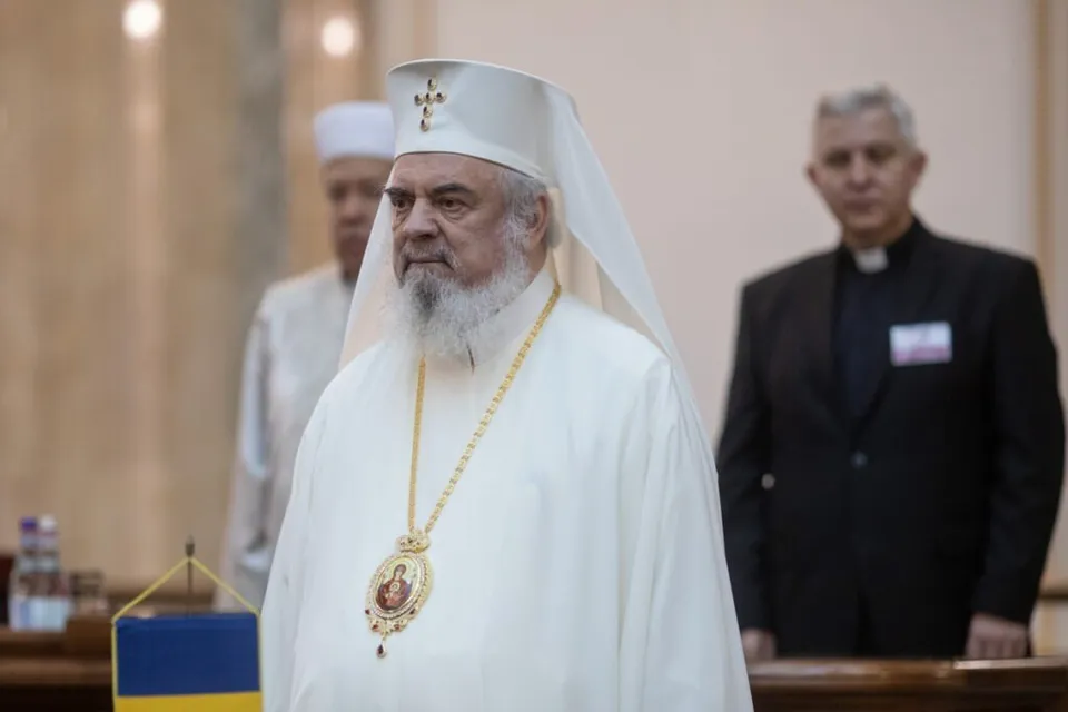 Un bărbat cu barbă albă poartă o vestimentație albă și o pălărie albă cu o cruce, având un medalion suspendat la gât.