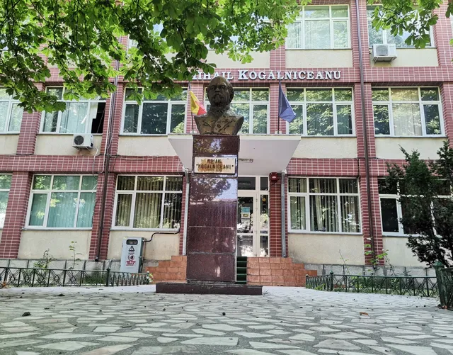 O bustă de bronz este așezată pe un soclu înalt în fața unei clădiri de cărămidă roșie cu ferestre mari și vegetație în jur.