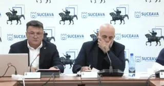 Un bărbat cu ochelari și sacou închis stă la o masă lângă un bărbat chel, care are mâna la față, în fața unui fundal alb cu logouri.