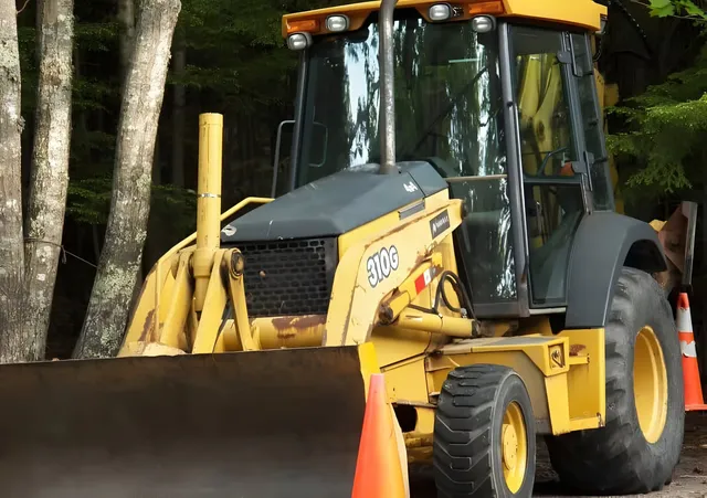Un buldoexcavator galben cu o cupă frontală este parcat lângă câțiva arbori, lângă un con de circulație portocaliu.