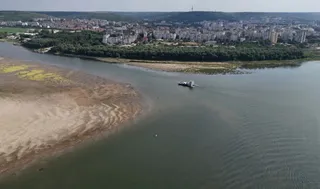 O navă mică navighează pe un fluviu larg lângă o zonă de nisip expusă și o rive acoperită de vegetație verde.