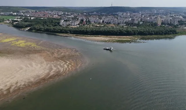 O navă mică navighează pe un fluviu larg lângă o zonă de nisip expusă și o rive acoperită de vegetație verde.