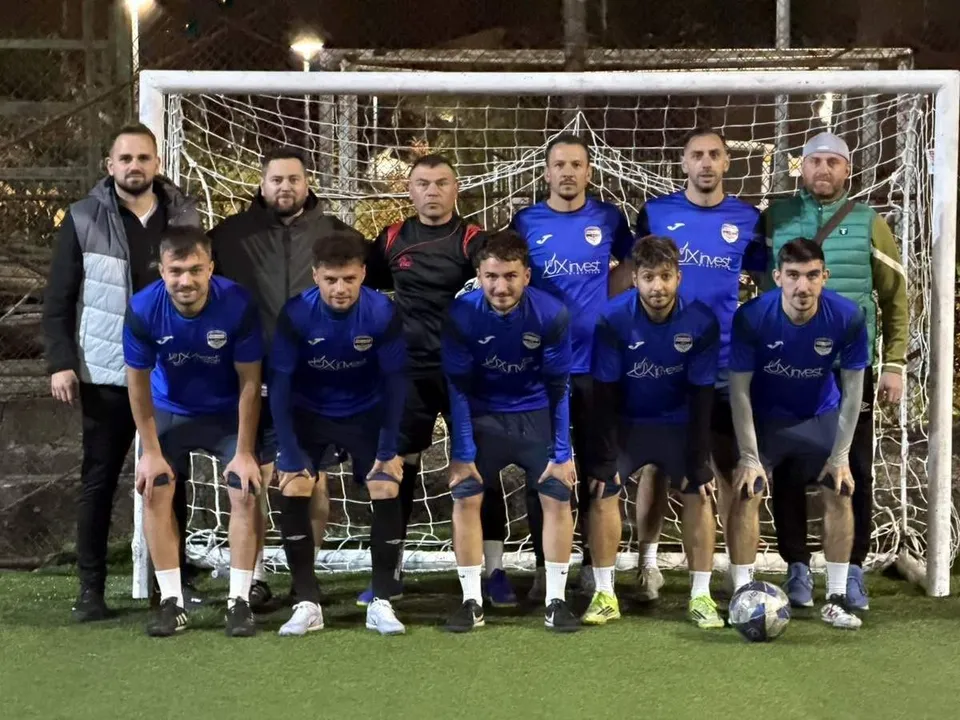 O echipă de nouă bărbați în echipamente sportive albastre și negre pozează în fața unei porți de fotbal pe un teren sintetic noaptea.