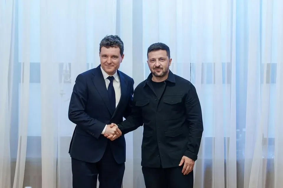 Doi bărbați stau în picioare în fața unor draperii albe și își strâng mâinile într-o strângere de mână.