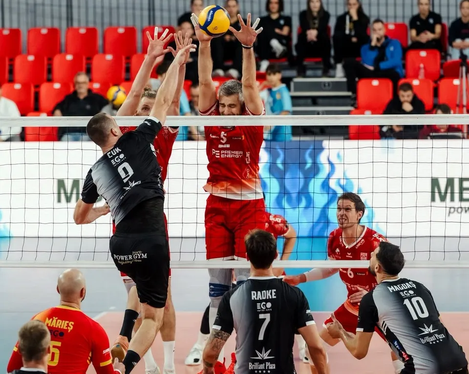 Câțiva jucători de volei de pe un teren de sport sar spre o minge galbenă și albastră deasupra unei nete întinse.