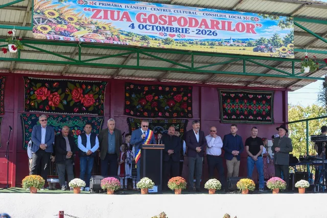 Un grup de oameni stă în picioare pe o scenă decorată cu tapiserii florale și un banner colorat sub un acoperiș metalic.