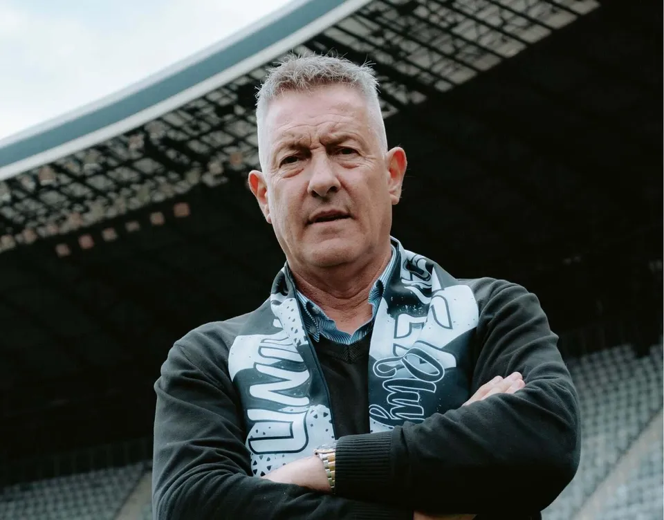 Un bărbat cu părul scurt și gri stă cu brațele încrucișate purtând un hanorac negru cu un model alb pe piept în fața unor tribune.
