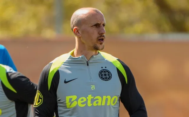 Un bărbat cu capul ras poartă un tricou sportiv gri cu accente galbene și negre, având un logo negru pe piept și textul Betano scris cu galben.