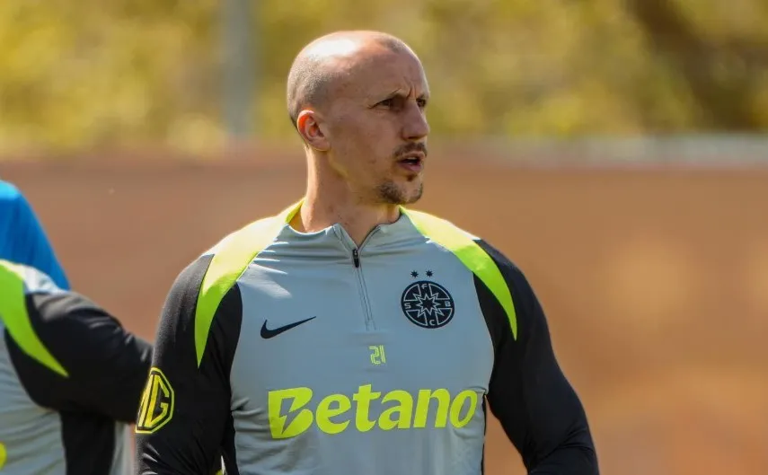 Un bărbat cu capul ras poartă un tricou sportiv gri cu accente galbene și negre, având un logo negru pe piept și textul Betano scris cu galben.