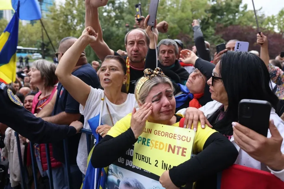 O femeie plânge în timp ce este îmbrățișată de alte persoane într-o mulțime care deține un afiș galben cu text negru și albastru.