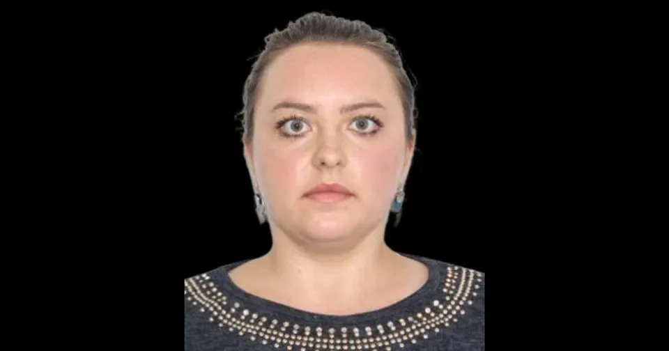 O femeie cu părul strâns în spate și cercei argintii poartă un tricou gri cu detalii metalice la guler pe un fundal negru.