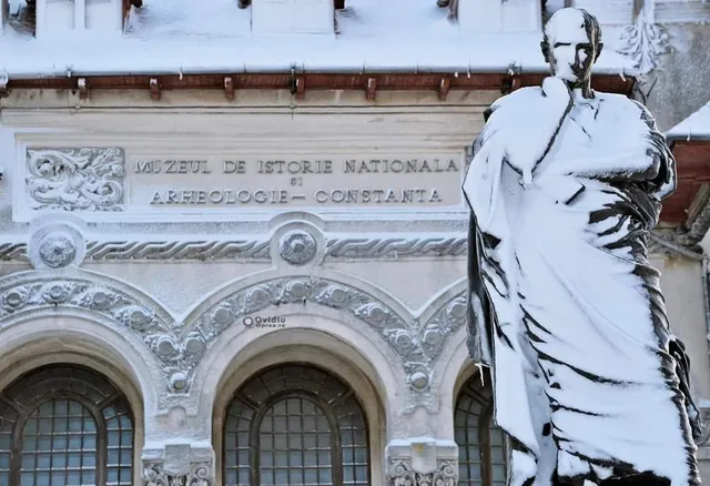 O statuie acoperită de zăpadă stă în prim-plan în fața unei clădiri albe cu ferestre arcuite și inscripție în relief.