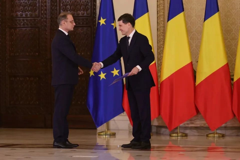 Doi bărbați în costume închise stau în picioare și se strâng în mână în fața unor steaguri într-o încăpere cu panouri de lemn sculptat.
