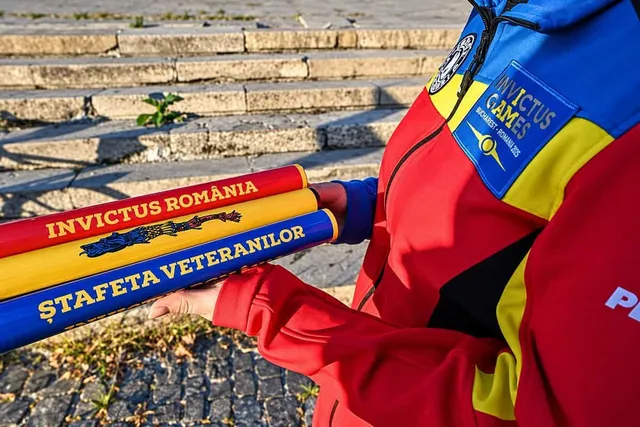O persoană poartă o jachetă colorată și ține în mâini un obiect cilindric tricolor cu inscripții textuale.