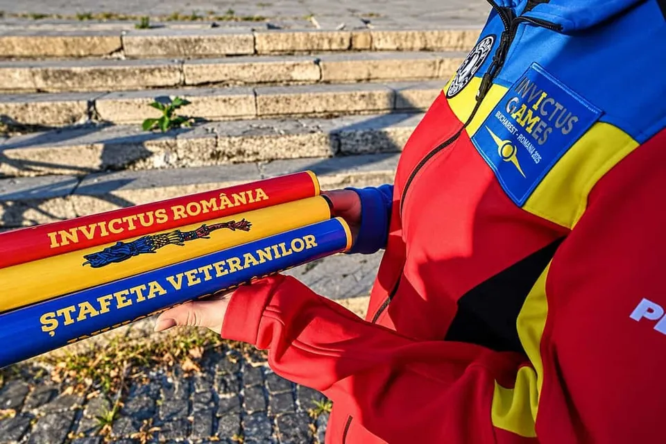 O persoană poartă o jachetă colorată și ține în mâini un obiect cilindric tricolor cu inscripții textuale.
