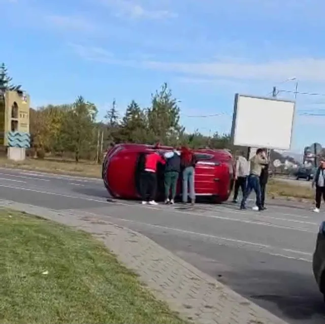 Un autoturism roșu este răsturnat pe lateral pe o stradă, în timp ce mai mulți oameni stau în picioare lângă vehicul.