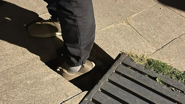 O persoană în pantofi deschiși și pantaloni închiși stă cu un picior într-o deschidere rectangulară a trotuarului de lângă o grilă metalică.