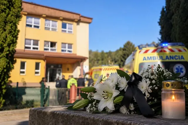 Un buchet de flori albe și o lumânare aprinsă sunt așezate pe un parapet, cu o clădire și o ambulanță în fundal.