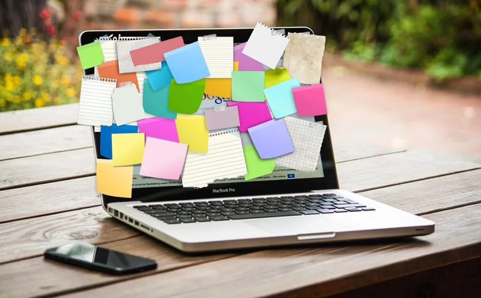 Un laptop deschis, acoperit cu note colorate, stă pe o masă de lemn lângă un telefon.