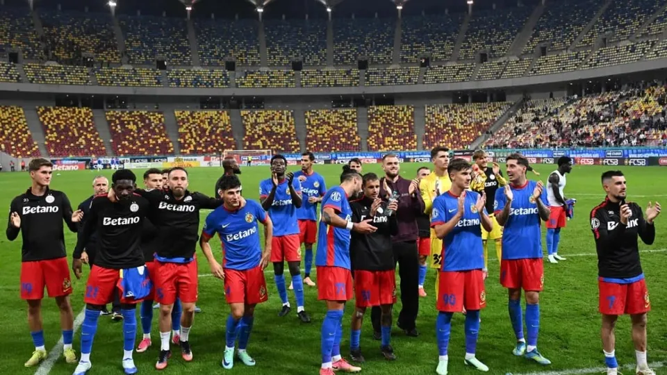 Un grup de fotbaliști în echipamente sportive albastre și negre stau pe un teren de fotbal verde în fața unor tribune pline de spectatori.